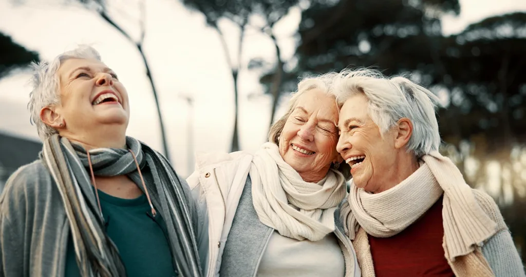 Three older adult women, laughing and smiling together as they walk through an outdoor park